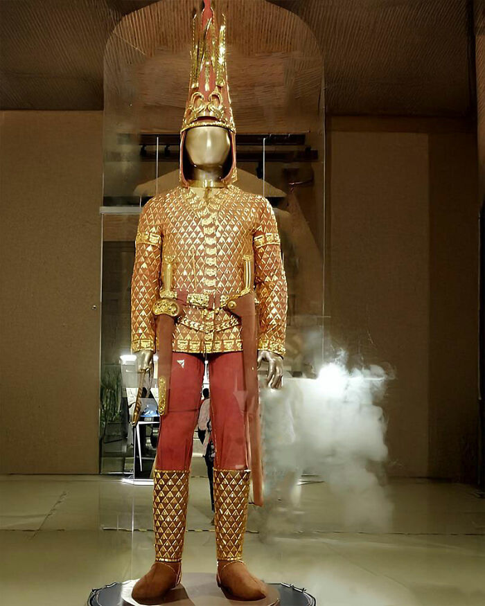 Ancient history gold ceremonial armor and helmet displayed in a museum with smoke effects in the background.