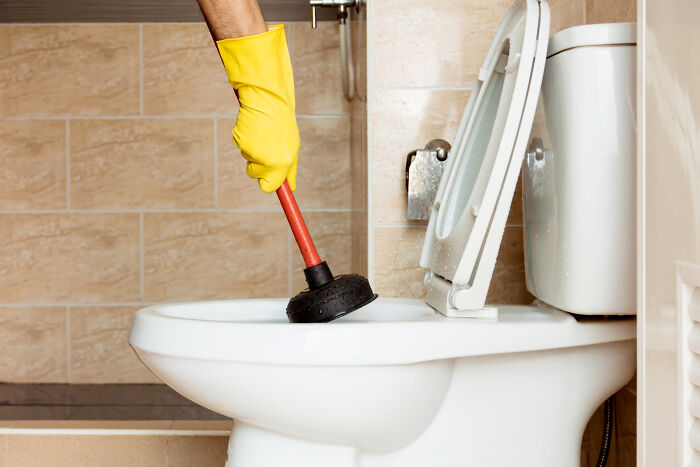 Person wearing yellow gloves using a plunger to fix a clogged toilet, depicting life blunders and secrets.