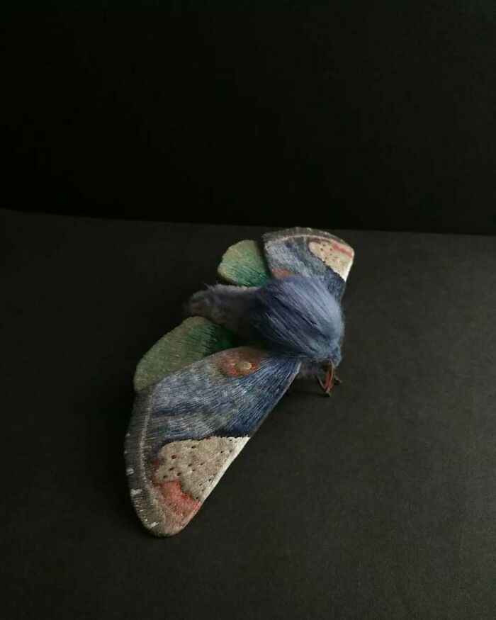 Embroidered fiber moth in shades of blue and green with detailed stitching on black background.