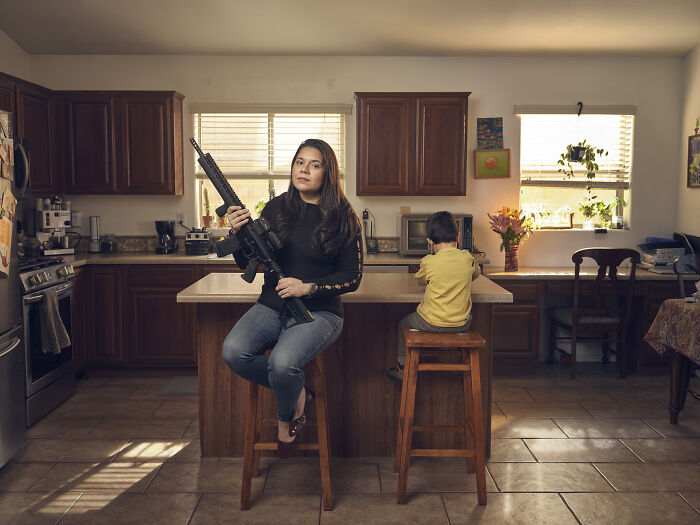 Woman holding a rifle sitting on a stool in a kitchen with a child beside her, capturing the essence of humanity.