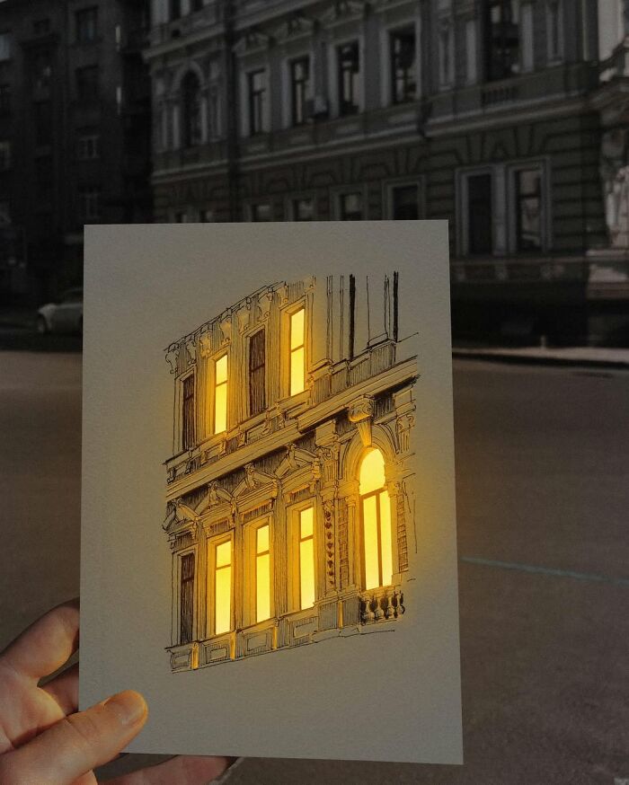 Pen and ink drawing of a building with glowing windows held against a real urban background, showcasing Ukrainian artist's work.
