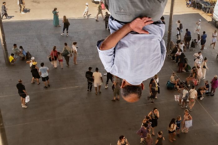 Man reflected upside down in a large mirror above a busy crowd in an urban setting, showcasing street photography.