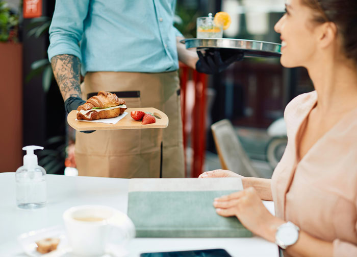 Waiter serving a croissant and strawberries to a seated woman, illustrating infuriating things said by people in charge.