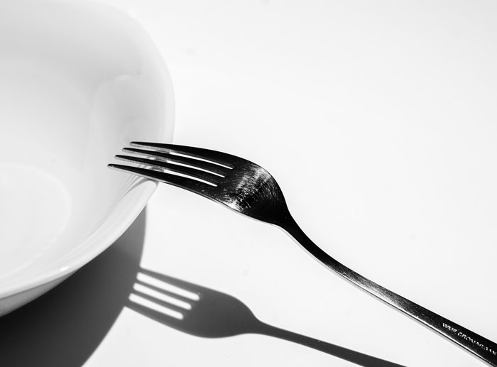 Fork resting on the edge of an empty white bowl casting a shadow, illustrating a weird habit related to being raised poor.