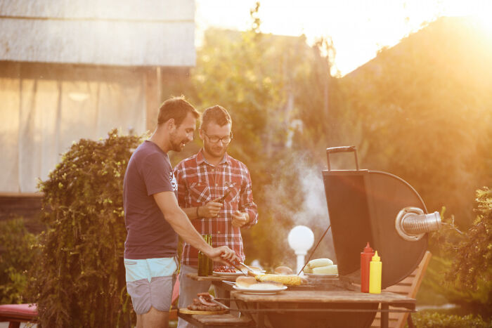Two men grilling outdoors in a messy garage setting with a refrigerator visible in the background at sunset.