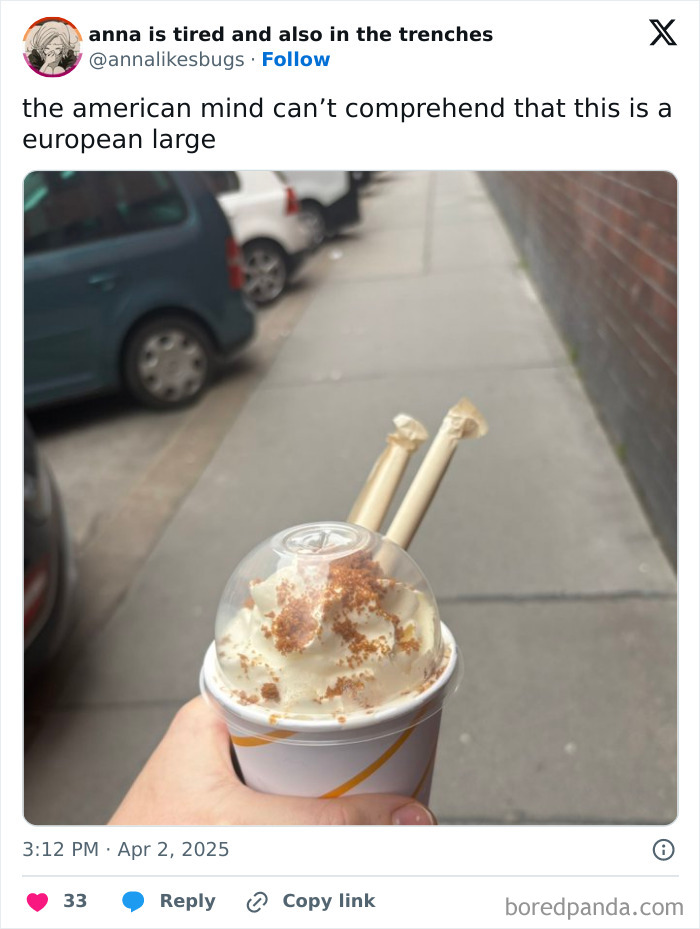 Hand holding a European large cup with whipped cream and two wrapped straws on a city sidewalk, confusing Americans.