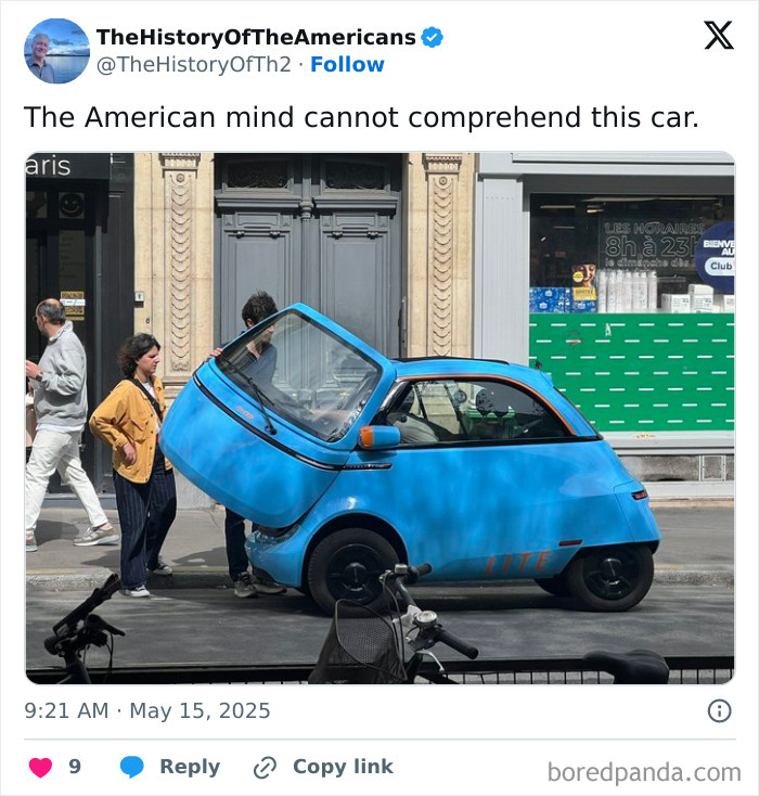 Small blue three-wheeled car with front door open on city street, capturing the American mind confusion concept.