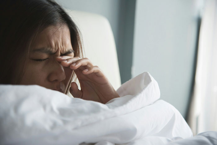 Woman in bed rubbing eyes, appearing tired or unwell, suggesting a scenario related to calling in sick.