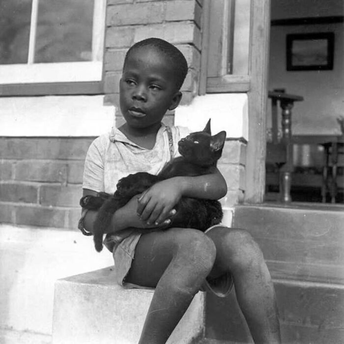 Vintage black and white photo of a child sitting outside, gently holding a black cat, showing the timeless bond between kids and cats.