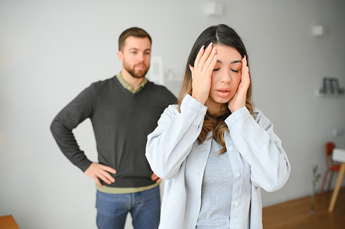 Woman holding her head in distress while man stands behind with hands on hips in a tense home setting. Woman holding her head in distress while man stands behind with hands on hips in a tense home setting.