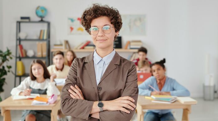 Confident young teacher with glasses in classroom, symbolizing understanding of dark family secrets shared by adults.