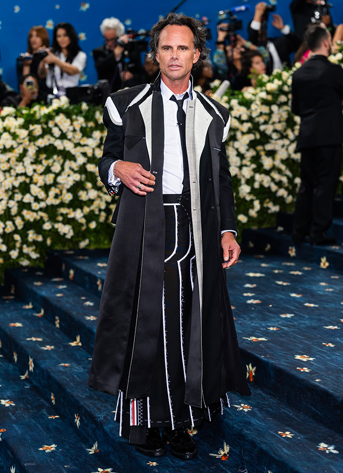Walton Goggins in a black and white outfit posing on a blue carpet with floral decorations at an event. Walton Goggins in a black and white outfit posing on a blue carpet with floral decorations at an event.