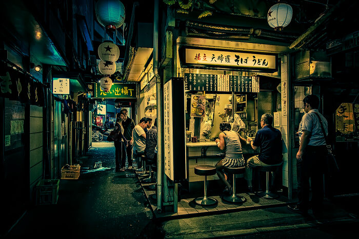 Night street photography moment showing people dining at a small urban eatery with glowing lanterns and signs.
