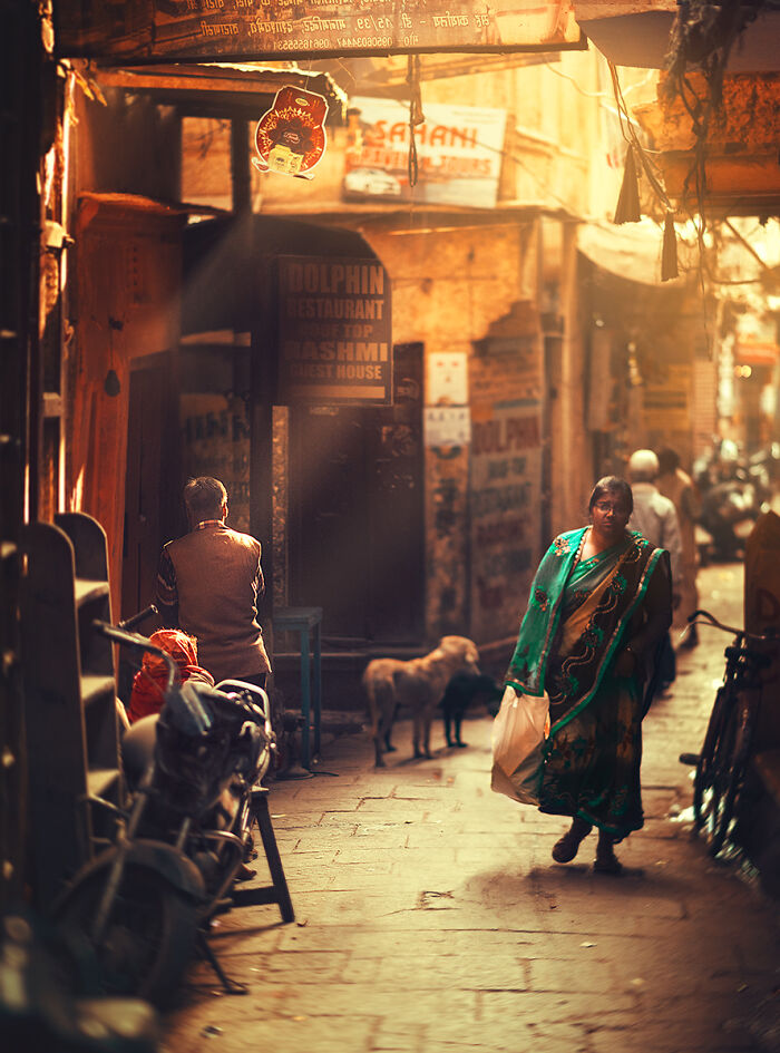 Street photography moment of a woman walking through a sunlit narrow alley with people and animals in urban setting.