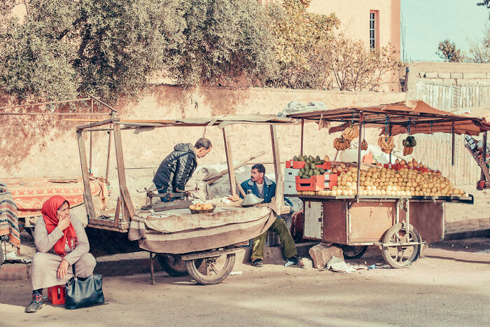 Street photography moment showing market vendors with fruit stands and a woman sitting nearby in an outdoor setting.