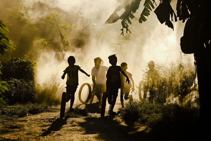 Children running on a dirt path surrounded by smoke and foliage in a vibrant street photography moment.