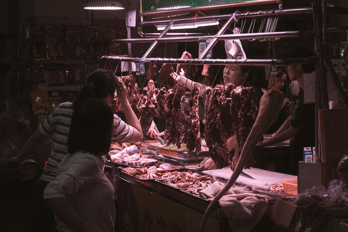 Street photography moment showing a busy night market vendor selling hanging meats to multiple customers.