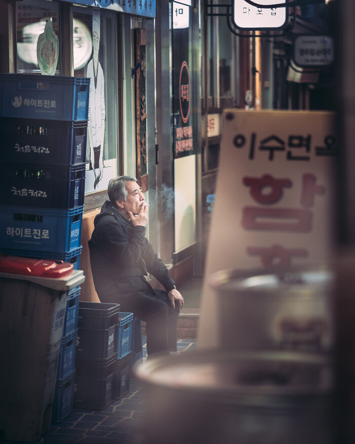 Man smoking outside a shop in an urban setting, a quiet street photography moment captured by talented photographers.