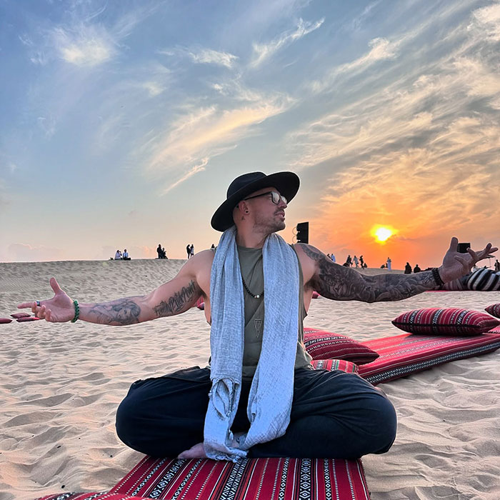 Man with tattoos and hat meditating at sunset on desert sand, reflecting former pole dancer turned monk story.