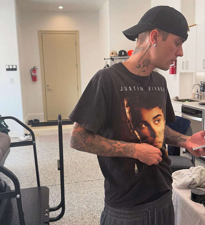 Justin Bieber wearing a black cap and graphic t-shirt indoors, checking his phone, showing tattoos on his arms and neck.