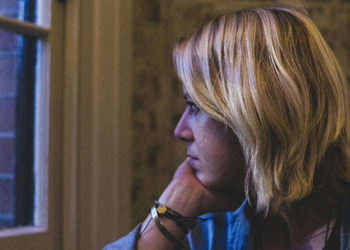 Woman with blonde hair looking thoughtfully out a window, reflecting on neighbor stealing packages and getting petty.