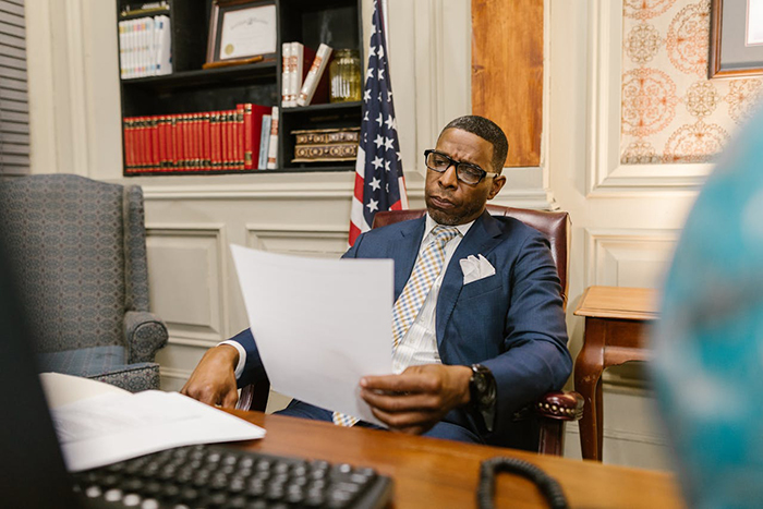 Man in suit reading documents at desk with American flag, related to cop wrongfully tickets driver for speeding case Man in suit reading documents at desk with American flag, related to cop wrongfully tickets driver for speeding case