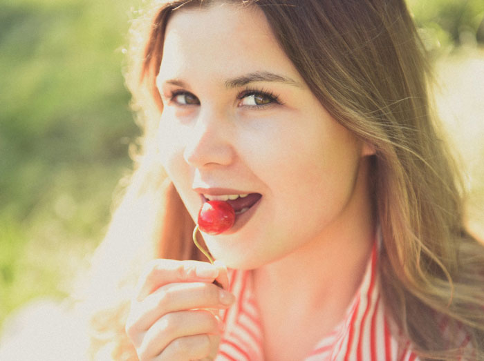 Young woman with long hair enjoying a cherry outdoors, representing problematic behaviors women get a pass for according to men.