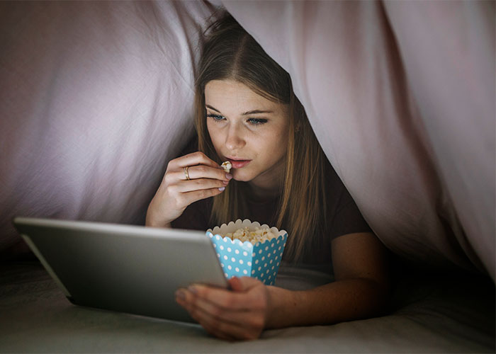 Young woman enjoying popcorn while watching a tablet under a blanket, highlighting things everyone does but doesn’t talk about.