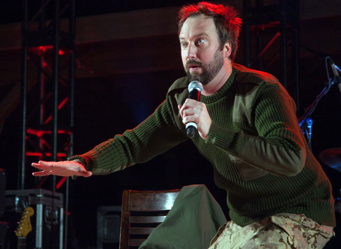 Comedian in green sweater holding microphone during a live celebrity encounter event on stage with dramatic lighting.