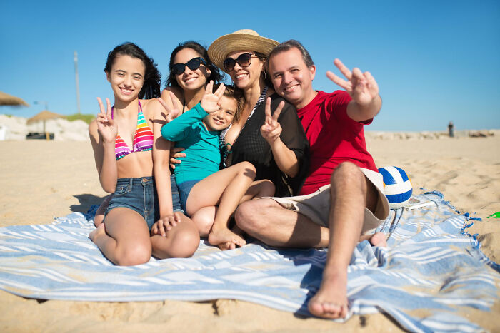 A happy family of five sitting on a beach blanket, smiling and making peace signs on a sunny day.