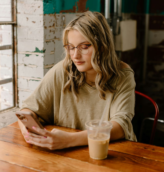 Young woman with glasses sitting indoors, looking at her phone, reflecting on a friendship of 10+ years ended.