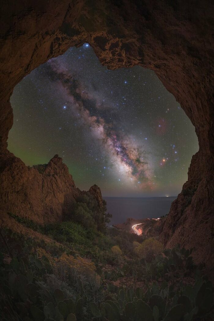 Milky Way shot framed by rocky cave opening overlooking starry night sky and distant ocean coastline.
