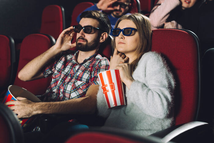 Couple wearing 3D glasses at a movie theater, holding popcorn, illustrating people name everyday items that are expensive.