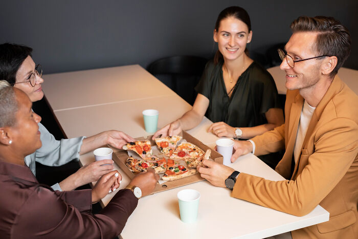 Four diverse adults sharing pizza while smiling, capturing moments from adult life that feel like being picked last in gym class.