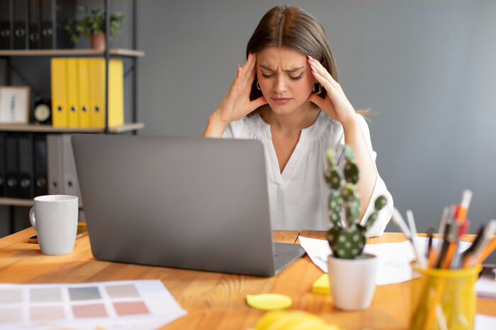 Young woman with headache at office desk using laptop, illustrating casual things that can actually be deadly risks.