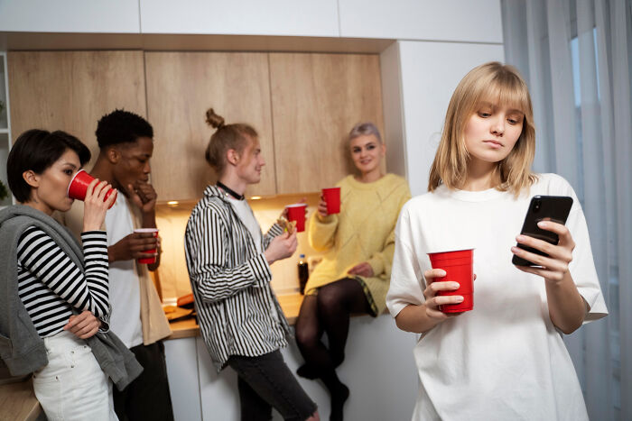 A young woman looking at her phone feeling excluded while others socialize, illustrating adult life moments like being picked last.
