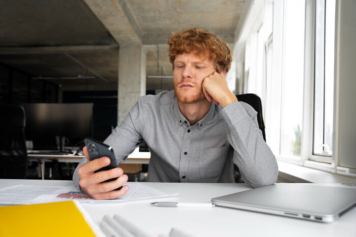 Young man looking disappointed while checking phone at office desk, capturing moments of feeling picked last in gym class.
