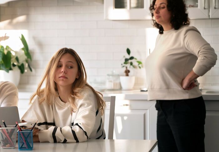 Teen girl looking upset at kitchen table while mother stands with arms on hips, illustrating soul-crushing things parents told kids.