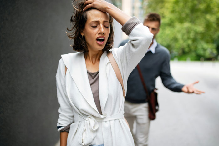 Woman showing problematic behaviors while man in background appears frustrated during an outdoor conversation.