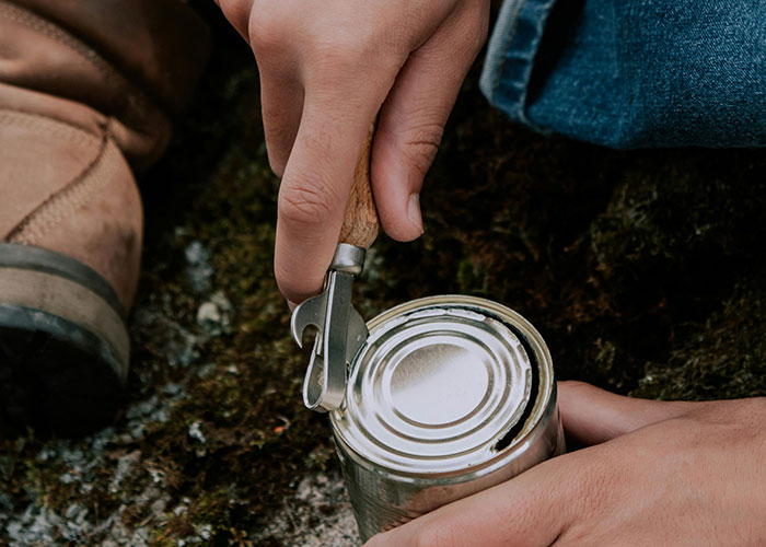 Person using a manual can opener to open a tin can outdoors with boots and moss visible in the background for entertaining facts.