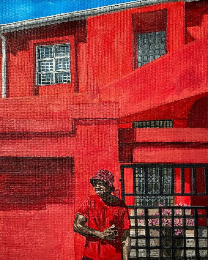 Man in red shirt and hat standing in front of a vivid red building, highlighting colours, space, and identity in the painting.