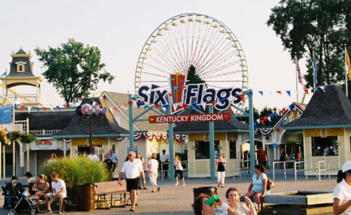 Visitors walking at Six Flags Kentucky Kingdom amusement park with a Ferris wheel, symbolizing former employees revealing workplace secrets.