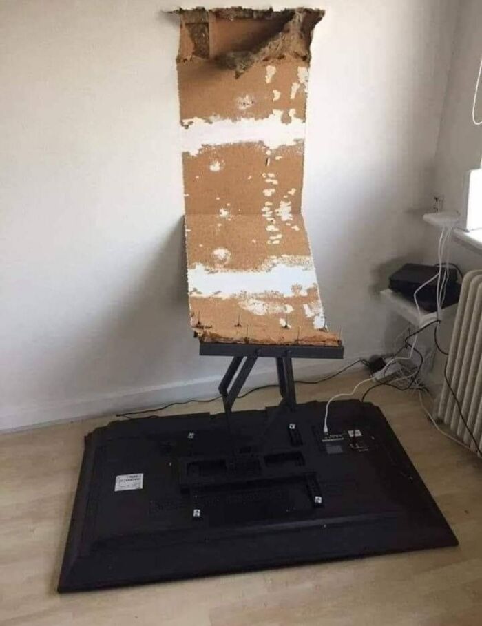 Failed renovation showing a broken wall patched poorly with cardboard leaning on a fallen TV on the floor.