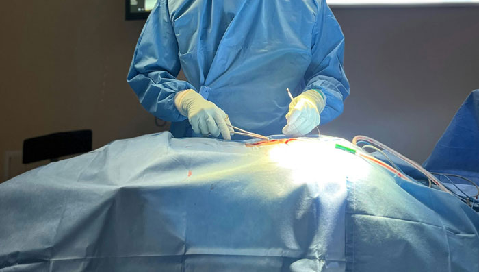 Experto en autopsias con guantes y bata azul realizando un procedimiento quirúrgico en sala médica iluminada.