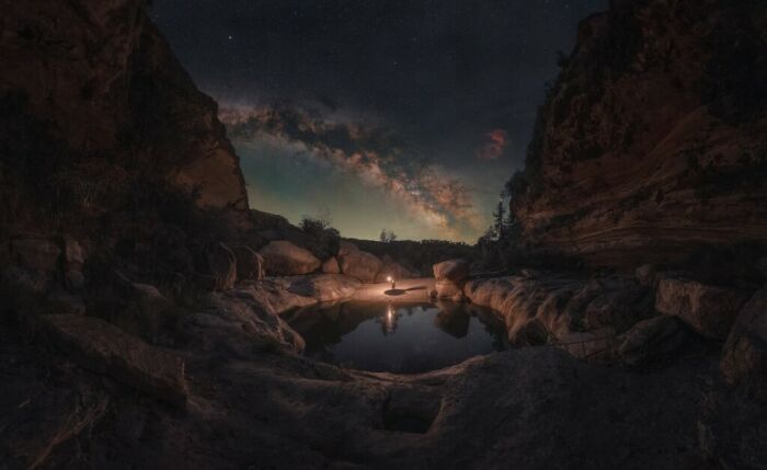 Person sitting by a reflective water pool in a canyon under a stunning Milky Way night sky showcasing natural beauty.