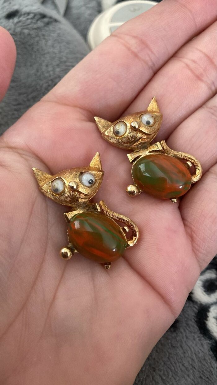 Pair of gold-tone thrifted cat brooches with green and orange stone bodies held in a hand, showcasing weird and wonderful thrifted finds.