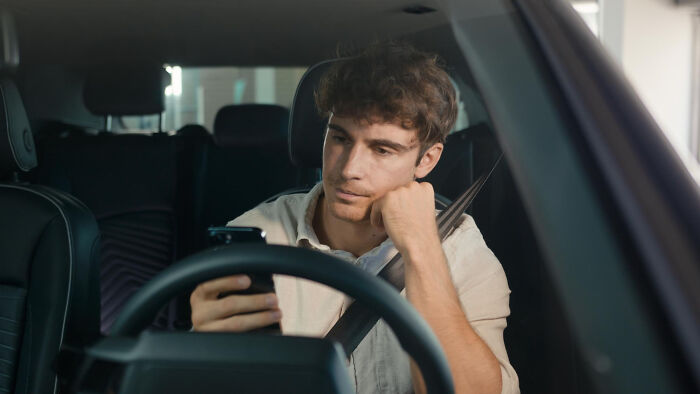 Young man sitting in a car looking bored at his phone, representing adult life moments feeling like being picked last.