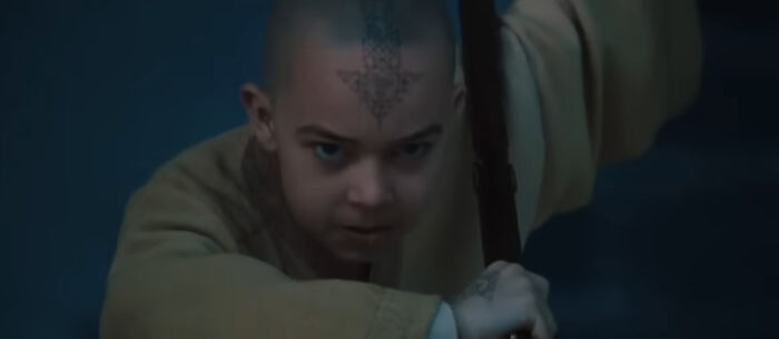 Young miscast actor with a serious expression holding a staff, portraying a tense movie scene in dim lighting.