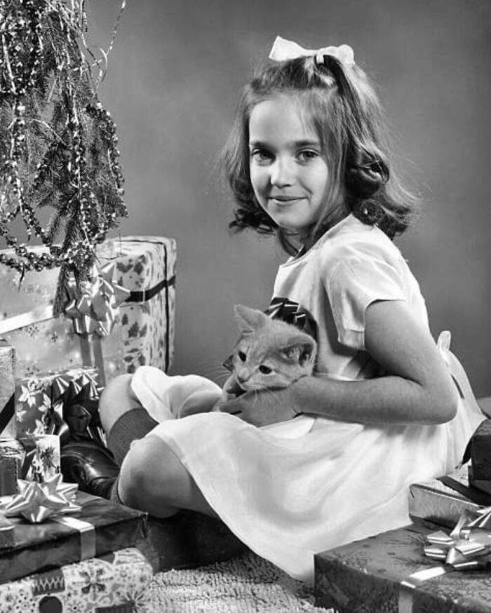 Vintage photo of a young girl holding a cat surrounded by Christmas presents, showcasing the timeless bond between kids and cats.