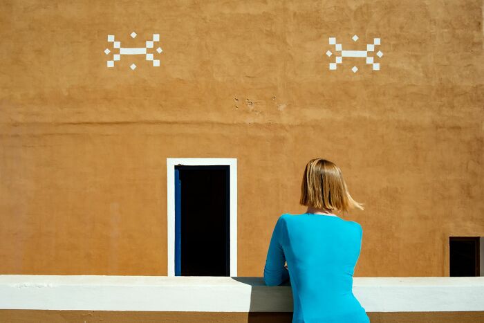 Woman in a bright blue top looking at a textured orange wall with dark doorways, capturing a street photography moment.